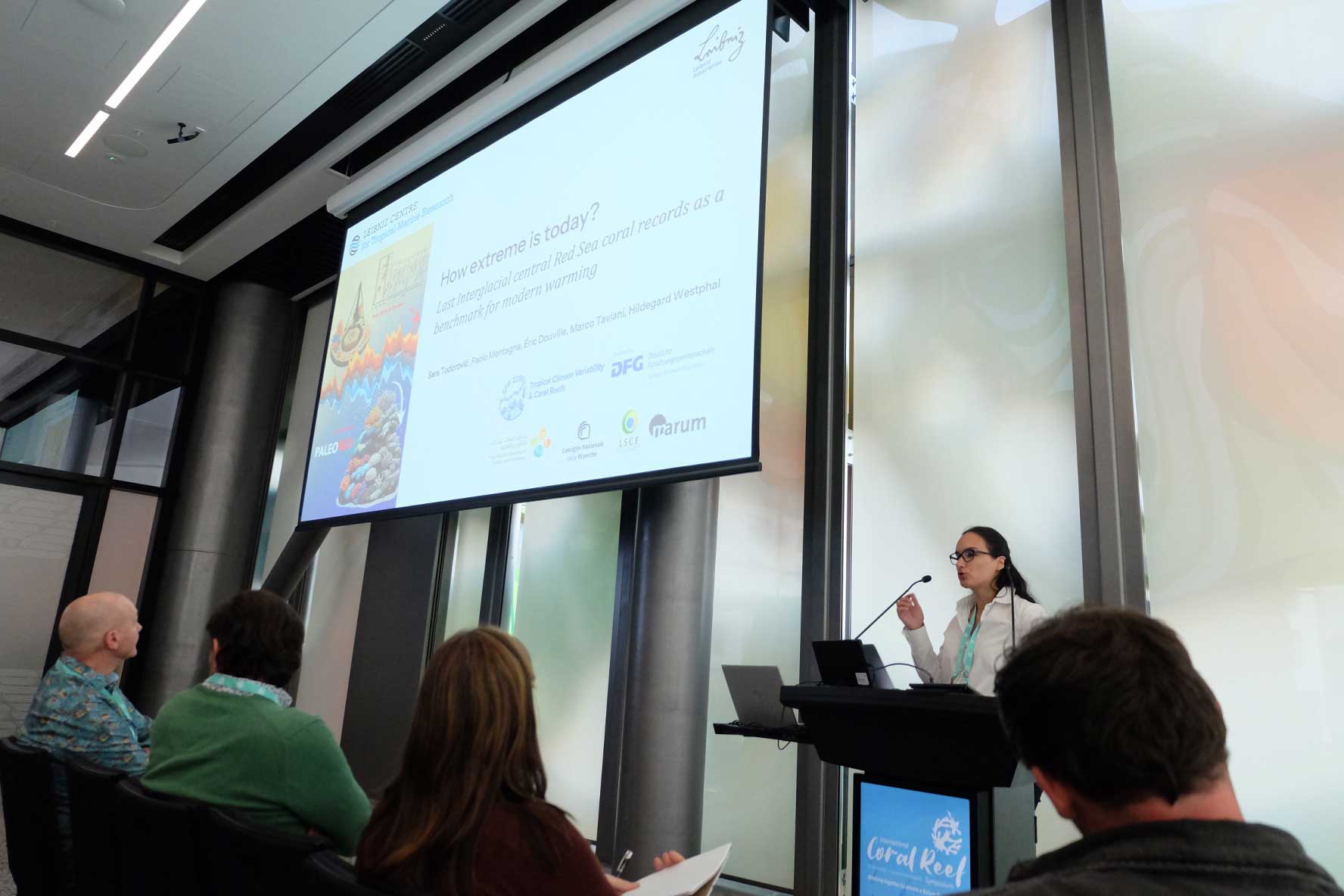 A woman gives a presentation to an audience about coral reefs and climate data. A large screen above her displays charts and the title “How extreme is today?”.