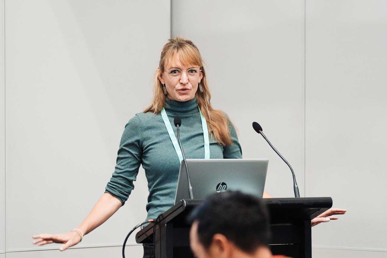 A woman gives a presentation at a lectern equipped with a laptop and microphones. Part of the audience is visible out of focus in the foreground.