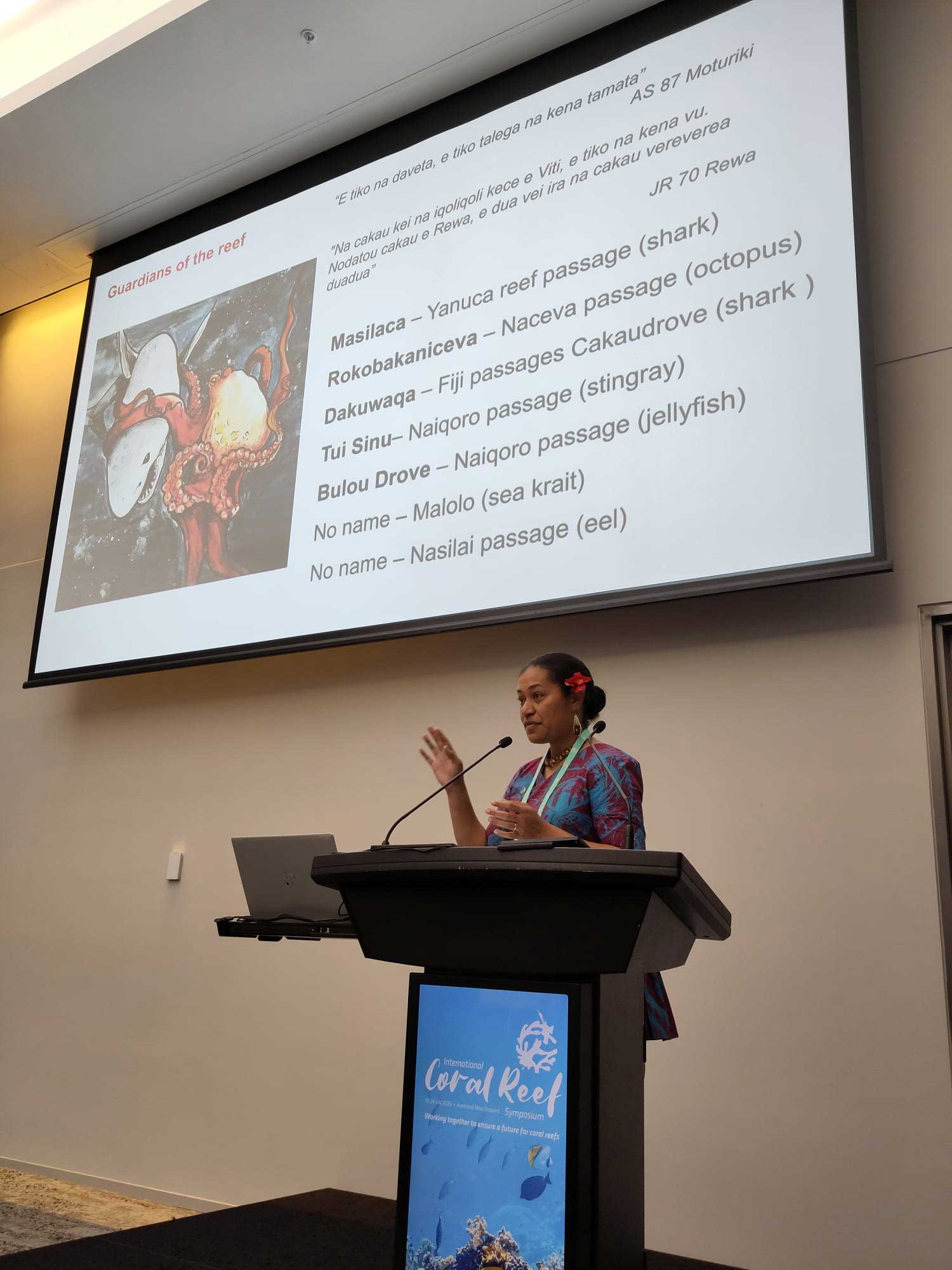 A woman gestures while speaking at a lectern. The large presentation screen behind her shows a slide titled “Guardians of the reef,” listing various marine animals and reef passages.