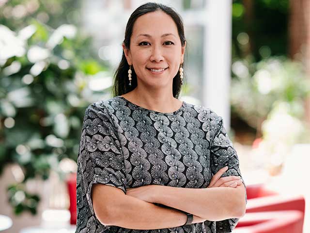 A woman stands confidently with her arms crossed, smiling at the camera. She has long dark hair pulled back and is wearing dangling earrings and a patterned black and gray top with circular designs. The background is softly blurred, showing green plants and a bright, airy indoor or patio setting with natural light.
