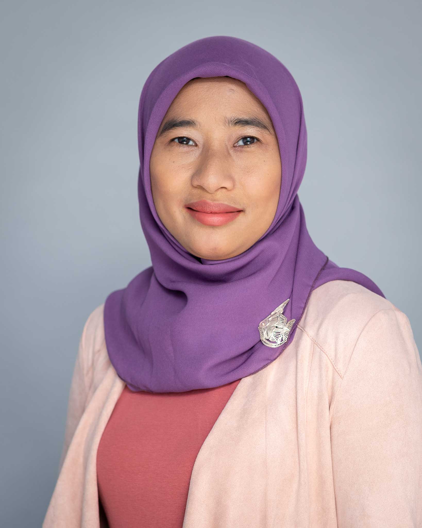 Protrait photo of a women with purple head scarf and a light pinkblazerin front of a grey background.