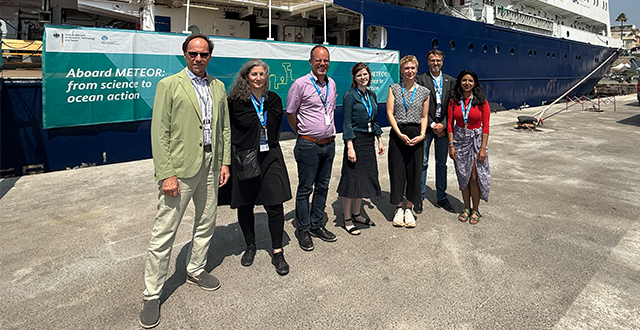 Seven people stand side by side on a dock in front of a large research vessel with a banner that reads "Aboard METEOR: from science to ocean action." They are dressed in business casual clothing and wearing lanyards. The ship behind them has a dark blue hull with white upper decks. The group is smiling and posing for the photo under bright sunlight.