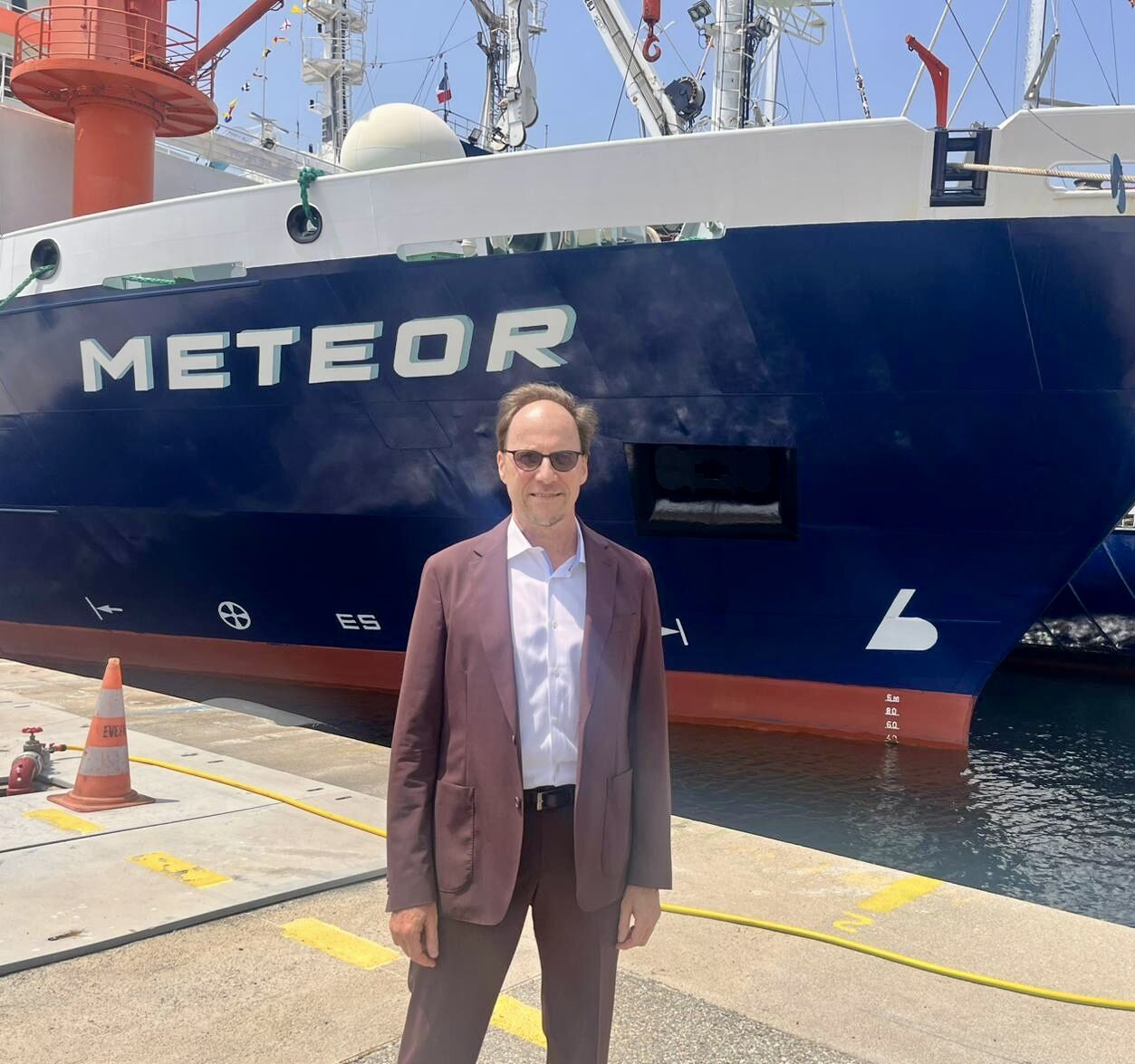 Alt-text: A man wearing a maroon suit and sunglasses stands on a dock in front of a large research vessel named "METEOR." The ship has a dark blue hull with white lettering and red accents near the waterline. Behind the man, part of the ship's equipment and antennas are visible on the upper deck. The scene is brightly lit under a clear, sunny sky, and an orange traffic cone is placed on the dock nearby.