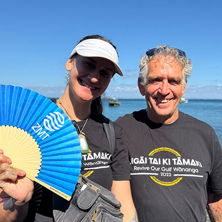 The photo shows a woman and a man in grey T-shirts in the sunshine against a blue sky. The woman is holding a blue paper fan labelled ZMT up to the camera.