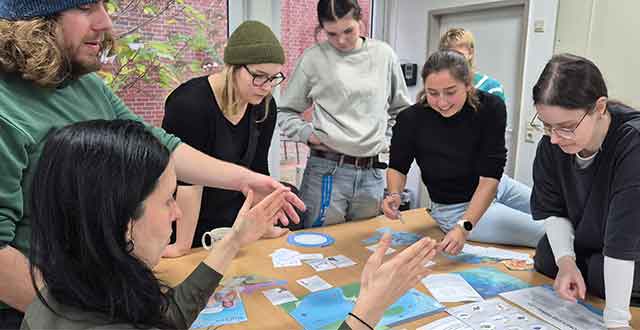 Sechs Erwachsene stehen und sitzen um einen Tisch, der mit blauen Karten, ausgedruckten Blättern und kreisförmigen Diagrammen bedeckt ist. Eine Person gestikuliert mit beiden Händen, während die anderen sich über den Tisch beugen und die Materialien aufmerksam betrachten. Die Gruppe wirkt engagiert in einer gemeinsamen Diskussion oder Arbeitsphase in einem hellen Raum.