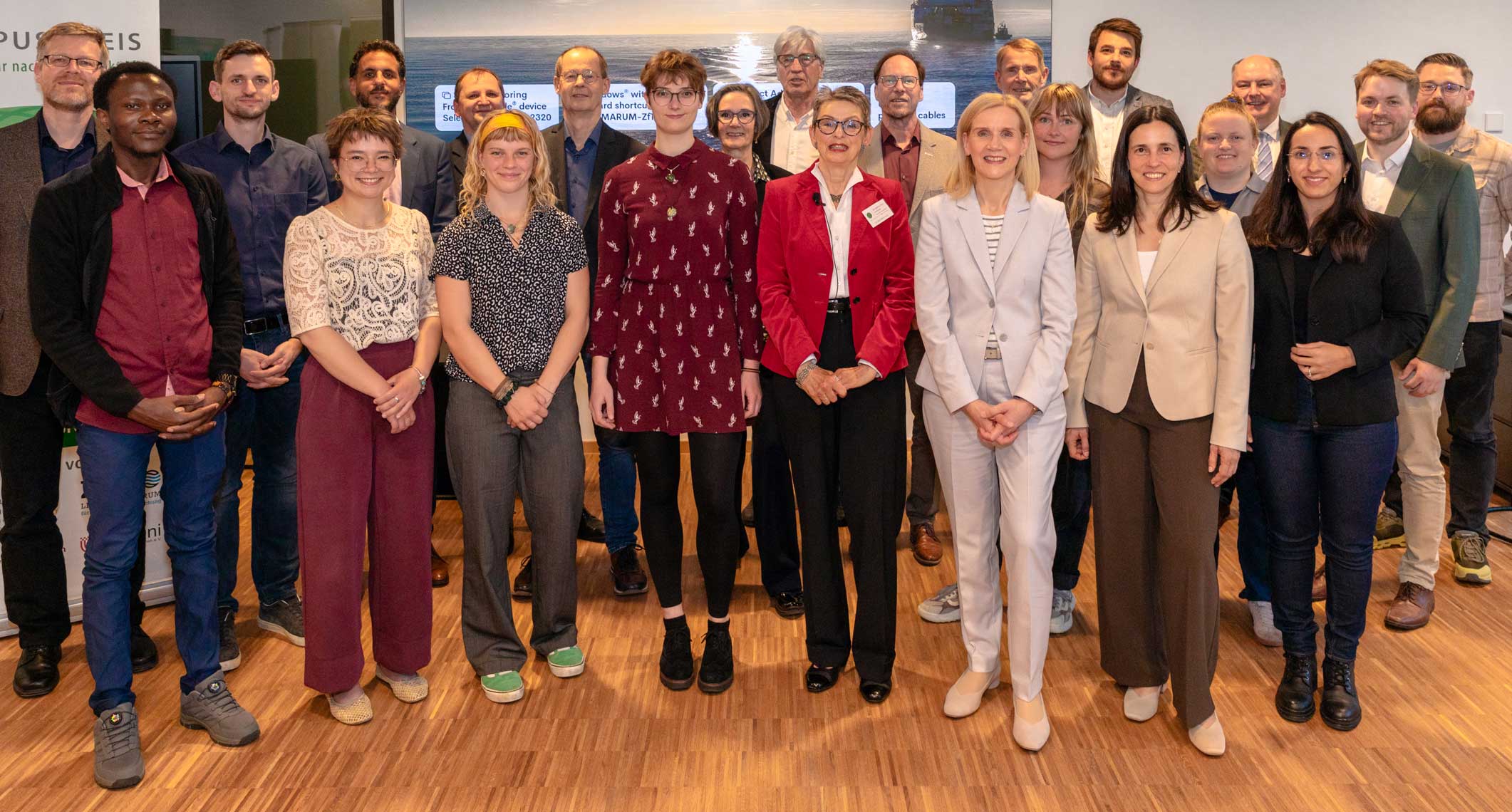 Großes Gruppenfoto von etwa zwanzig Forschern und Fachleuten, die in einem Innenraum auf einem Holzboden vor einer großen Leinwand stehen. Zu der Gruppe gehören Männer und Frauen, von denen einige in die Kamera lächeln. 