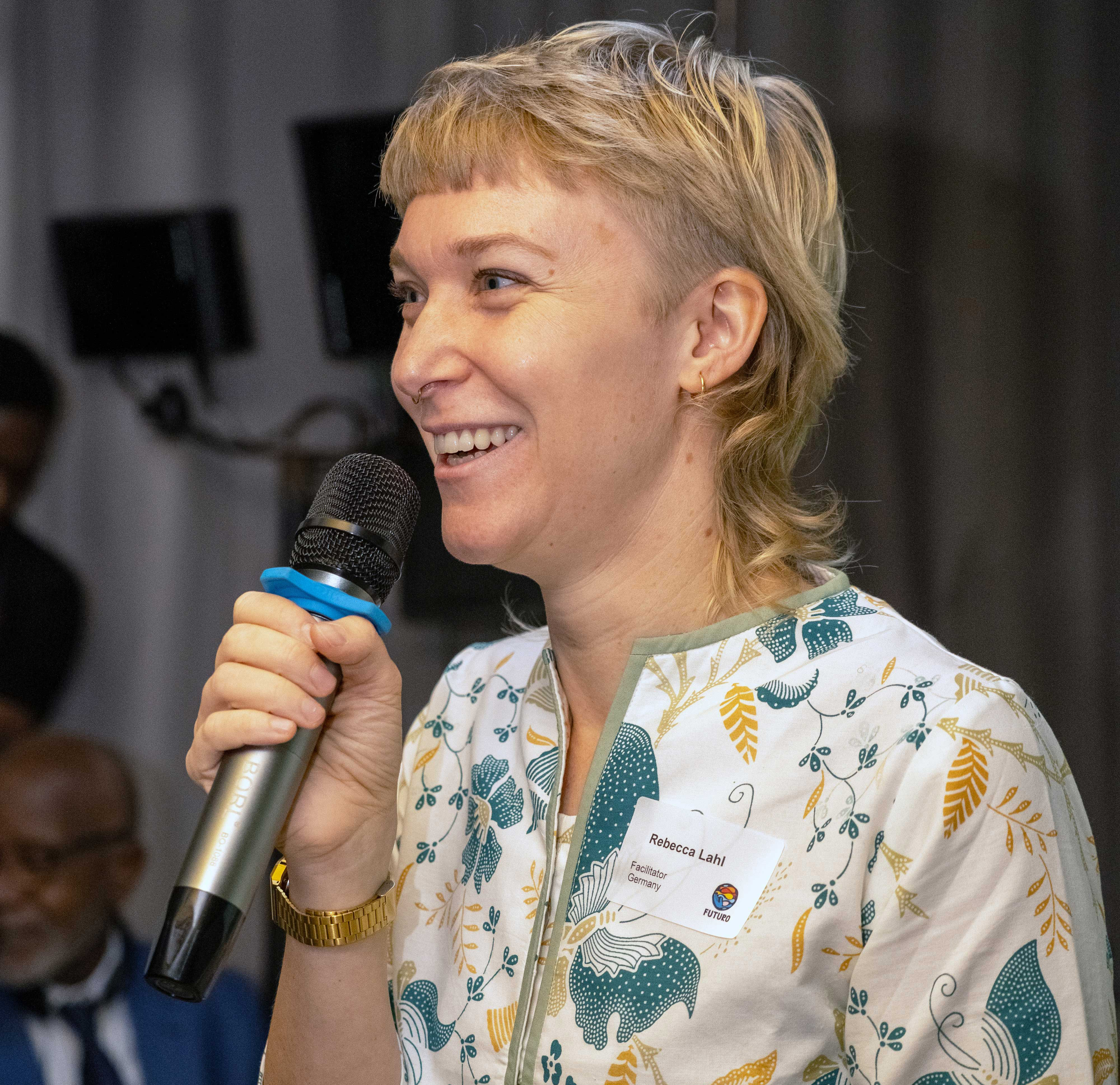 Eine lächelnde Frau mit kurz geschnittenem, blondem Haar hält ein Mikrofon in der Hand und spricht. Sie trägt eine weiße Bluse mit grün-gelben floralen Mustern und ein Namensschild mit der Aufschrift „Rebecca Lahl, Facilitator, Germany“ sowie dem Logo von FUTURO.