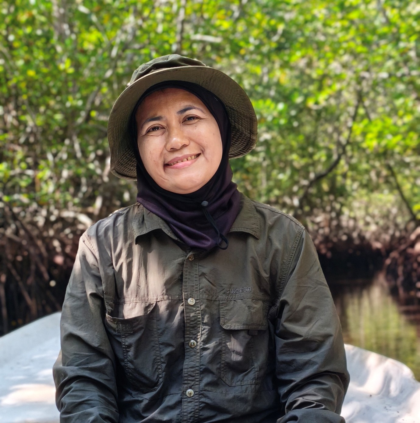 Portrait of a smiling woman wearing a hat in a lush mangrove landscape. Dense trees and a waterway are visible in the background.