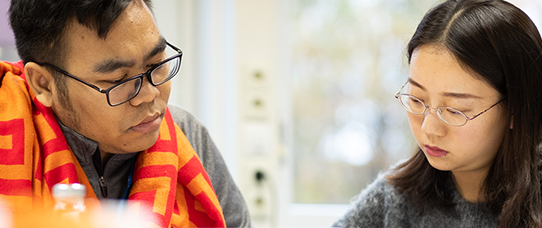 Alt-Text (Deutsch): Zwei Personen sitzen nebeneinander an einem Tisch und konzentrieren sich auf das Lesen oder Schreiben. Die Person links trägt eine Brille, eine graue Jacke und einen auffälligen rot-orange gemusterten Schal über den Schultern. Die Person rechts trägt ebenfalls eine Brille und einen grauen, flauschigen Pullover. Beide schauen nach unten und wirken vertieft