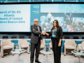 A man and a woman in business attire shake hands and smile while standing on a stage at a formal event. Behind them is a large screen displaying the event title, “Launch of the All-Atlantic Network of Coastal Resilience Beacon Sites.