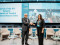 A man and a woman in business attire shake hands and smile while standing on a stage at a formal event. Behind them is a large screen displaying the event title, “Launch of the All-Atlantic Network of Coastal Resilience Beacon Sites.