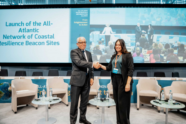 A man and a woman in business attire shake hands and smile while standing on a stage at a formal event. Behind them is a large screen displaying the event title, “Launch of the All-Atlantic Network of Coastal Resilience Beacon Sites.