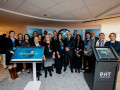 A diverse group of around 20 people stands together indoors, smiling for a group photo in front of a digital display and a globe graphic.