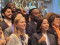 A large, diverse group of people stands closely together, smiling and celebrating at an indoor event. Many individuals are giving thumbs-up gestures and appear joyful and enthusiastic.