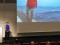 Vortragssituation in einem Konferenzsaal: Eine Referentin steht neben der Leinwand, auf der eine Aufnahme von einer Bootsfahrt auf dem Meer gezeigt wird.