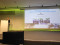 Blick auf die B&uuml;hne eines Konferenzsaals: Eine Referentin steht am Rednerpult neben einer gro&szlig; projizierten Pr&auml;sentationsfolie von Marion Glaser.