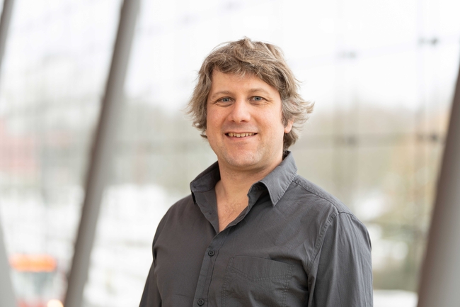 A smiling man with wavy, light brown hair and blue eyes stands indoors near large windows. He is wearing a dark grey button-up shirt with the top button open. The background is softly blurred, showing a bright, modern setting with tall glass panels and metal frames.