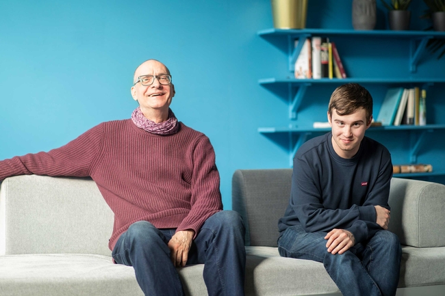 Two people are sitting on a light grey sofa: on the left, a man wearing glasses and a burgundy jumper; on the right, a teenager wearing a blue jumper and jeans. In the background, there is a blue wall and a bookshelf.