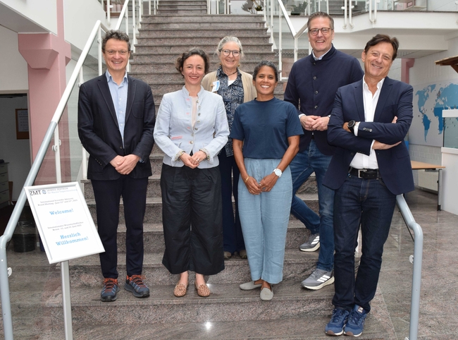 Six adults stand on a marble staircase indoors, posing for a group photo. Three men and three women are arranged in two rows, with the front row standing on a lower step. All are casually dressed in smart-casual attire, including blazers, button-up shirts, and relaxed pants. They are smiling and are in a professional or academic setting, with glass railings and a modern interior visible in the background.