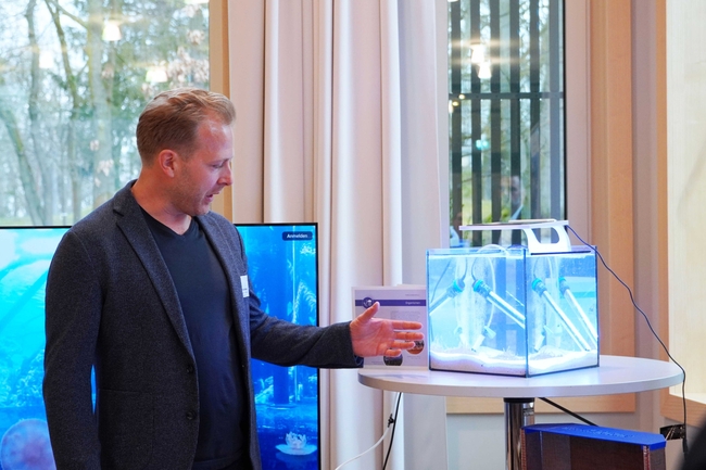 Ein Mann in dunklem Blazer und schwarzem Hemd gestikuliert in Richtung eines kleinen, hell erleuchteten Aquariums auf einem runden Tisch. Das Aquarium enthält weißen Sand, mehrere zylindrische Vorrichtungen mit Röhren und wird mit blauem Licht beleuchtet.