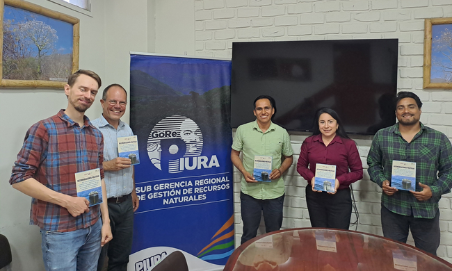 Five people stand indoors, holding copies of the same publication titled "Recomendaciones Políticas", which features a small boat on the ocean as the cover image. They are positioned in front of a banner displaying the logo of "GORE PIURA" and the text "Sub Gerencia Regional de Gestión de Recursos Naturales". Behind them is a brick wall with a large black screen and framed nature photographs.