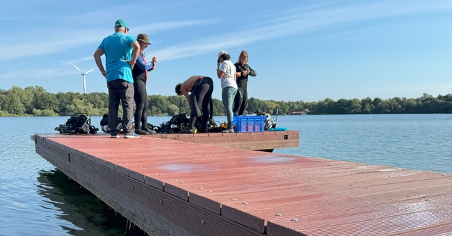 Several people are gathered on a wooden dock beside a clear lake, preparing for or assisting with scuba diving. Two divers in full black wetsuits and scuba gear sit on the edge of the dock, with one wearing green fins in the water. One person in a wetsuit stands behind them, helping adjust equipment. Three others in casual clothing stand or kneel nearby, holding or managing orange safety lines connected to the divers. The background features a tree-lined shore, a few wind turbines, and a partly cloudy blue sky.