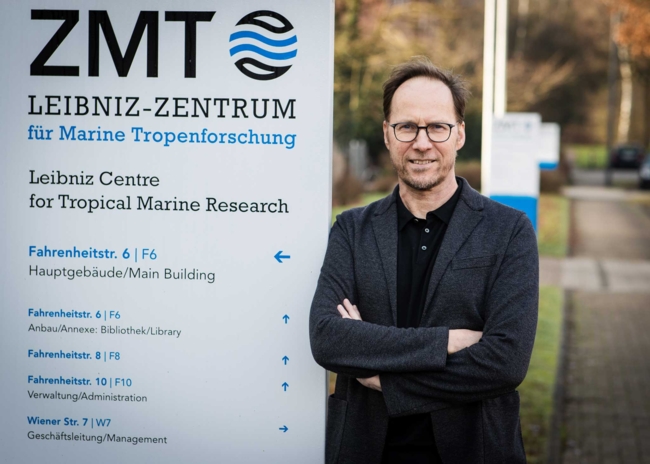 Mann in schwarzem jacket steht mit verschränkten Armen vor dem Schild des  Leibniz-Zentrums für Marine Tropenforschung (ZMT)