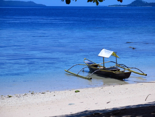 A small traditional outrigger boat with yellow supports and a white canopy is beached at the edge of a clear, shallow shoreline on a tropical island. The water transitions from light turquoise near the shore to deep blue further out.