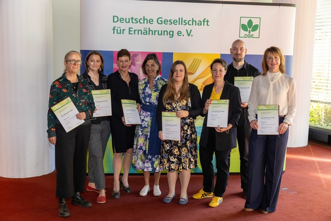 DGE Journalism Award Winners 2025 | Photo: DGE, Photo: TobiasVollmer.de Group photo of eight people standing indoors on a red carpet, holding certificates in front of a backdrop with the logo and name of the "Deutsche Gesellschaft für Ernährung e. V." (German Nutrition Society).
