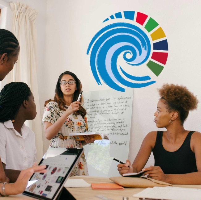 Vier Frauen befinden sich in einem hellen Raum und diskutieren gemeinsam. Eine Frau steht vor einem Whiteboard, hält einen Marker und ein aufgeschlagenes Buch in der Hand und spricht zu den anderen. Auf dem Whiteboard sind handschriftliche Notizen zu sehen. Zwei der Frauen sitzen am Tisch, hören aufmerksam zu, eine von ihnen macht sich Notizen. Im Vordergrund arbeitet eine weitere Frau an einem Tablet, auf dessen Bildschirm farbige Symbole zu erkennen sind.