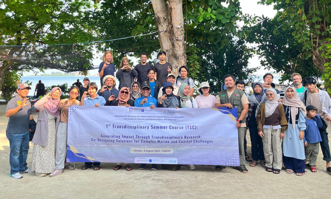 Eine große Gruppe von Menschen hat sich im Freien vor hohen Bäumen versammelt, im Hintergrund ist ein Stück Meer zu sehen. Sie lächeln und posieren gemeinsam hinter einem breiten Banner mit der Aufschrift: “1st Transdisciplinary Summer Course (TSC): Generating Impact Through Transdisciplinary Research: Co-Designing Solutions for Complex Marine and Coastal Challenges. 28 July – 8 August 2025 | Hybrid.”