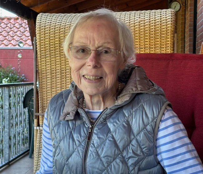 Eine Frau mit kurzen grauen Haaren und Brille sitzt auf einem Stuhl auf einer Terrasse und lächelt leicht in die Kamera. Sie trägt eine hellgraue, gesteppte Weste über einem blau-weiß gestreiften Shirt und eine Perlenkette. Hinter ihr steht ein Korbstuhl mit rotem Kissen; im Hintergrund sind ein Geländer, Pflanzen und Häuser mit roten Ziegeldächern im Tageslicht zu sehen.