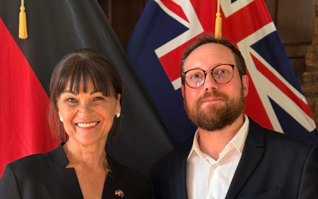 The photo shows a woman with dark hair in an elegant black jacket and a man with glasses, a beard and a black jacket with a white shirt standing in front of the New Zealandflag.