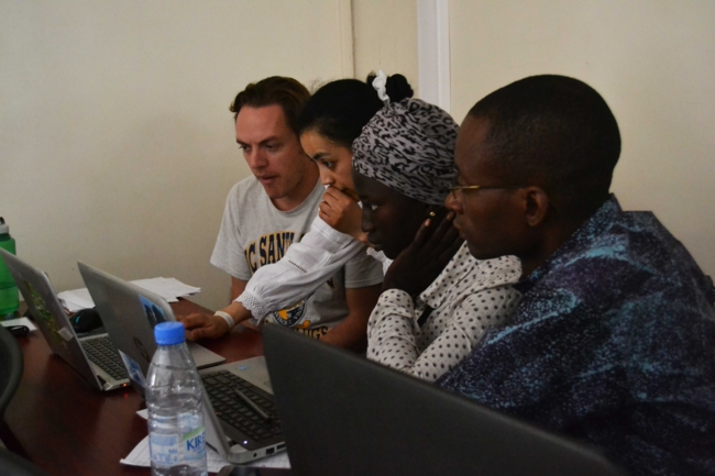 A group of four students sit in front of  their laptops, a female is pointing at the laptop screen