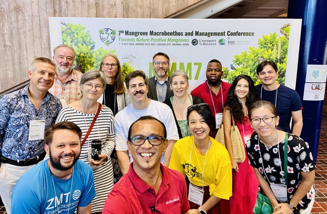 Gruppenfoto von Teilnehmenden der 7. Mangrove Macrobenthos and Management Conference vor einer Konferenzwand mit dem Motto „Towards Nature Positive Mangroves“. Die Gruppe blickt lächelnd in die Kamera und steht dicht beieinander. Im Hintergrund sind grafische Darstellungen von Mangroven sowie die Veranstaltungsinformationen zu sehen.