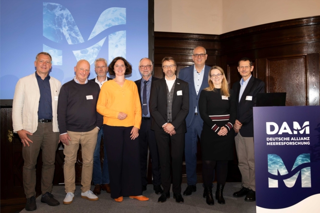 Gruppenbild von neun Personen auf eine Bühne, davon sind sieben Männer udn zwei Frauen, neben der Gruppe stehte ein Redepult mit der Aufschrift DAM - Deutsche Allianz Meeresforschung