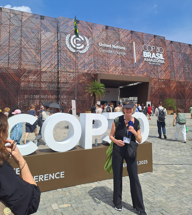 Eine Frau steht lächelnd vor einem großen „COP30“-Schriftzug am Eingang der COP30-Klimakonferenz. Sie trägt ein schwarzes Outfit, eine Kappe und ein Konferenz-Badge und hält ein Handy sowie ein Tuch in der Hand. Im Hintergrund ist eine moderne Fassade mit diagonalen Holzlamellen zu sehen, auf der die Logos der Vereinten Nationen zum Klimawandel sowie „COP30 Brasil Amazônia, Belém do Pará, 2025“ abgebildet sind. Über dem Eingang weht eine brasilianische Flagge, und zahlreiche Konferenzbesucher bewegen sich auf dem Platz bei sonnigem, blauem Himmel.