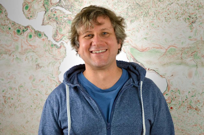 A smiling man with wavy, light brown hair stands in front of a colorful abstract map. He is wearing a blue zip-up hoodie over a blue t-shirt. The map behind him features swirling green, red, and beige patterns resembling topographical or oceanographic data.