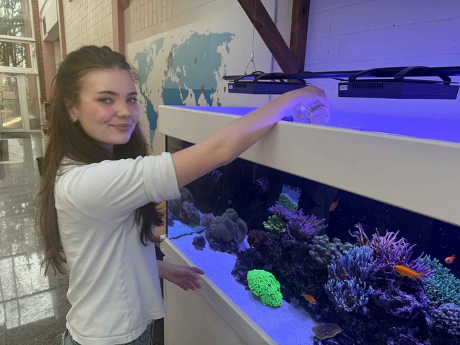 Frau mit langen braunen Haaren steht neben einem großen Meerwasseraquarium und lächelt in die Kamera, während sie Futter aus einem kleinen Behälter in das Becken gießt. Das Aquarium ist blau beleuchtet und enthält bunte Korallen sowie kleine orangefarbene Fische.