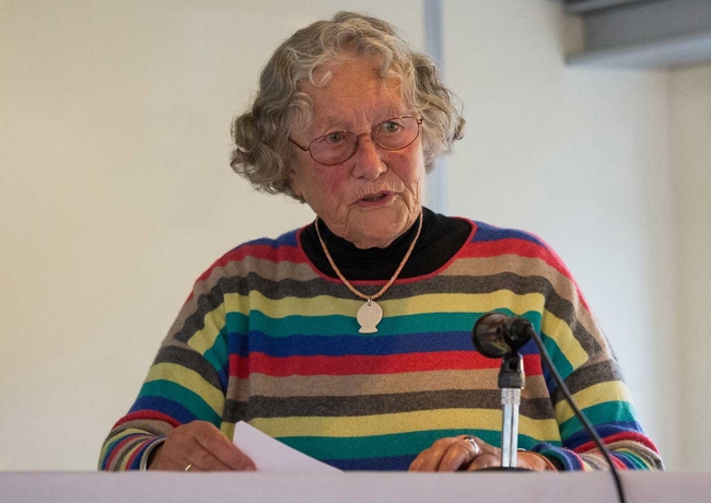 Eine Frau mit kurzen, lockigen grauen Haaren und Brille spricht an einem Rednerpult mit Mikrofon. Sie trägt einen bunten Pullover mit horizontalen Streifen in Rot, Gelb, Grün, Blau und Grau über einem schwarzen Rollkragenpullover. An einer Kette hängt ein Anhänger in Form eines Fisches. Die Frau hält ein Blatt Papier, während sie in einem weich beleuchteten Innenraum spricht.