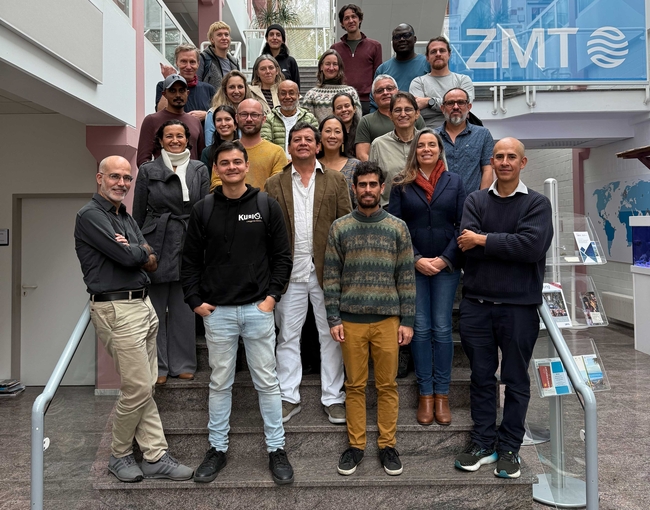 Eine Gruppe von Personen posiert für ein Foto auf einer Innentreppe in einem modernen Gebäude mit hohen Decken und Oberlichtern. Die Gruppe ist in mehreren Reihen auf den Stufen angeordnet, lächelt und blickt in die Kamera. Die meisten sind in Freizeit- oder Business-Casual-Kleidung gekleidet. Rechts im Bild steht ein großes blaues Banner mit dem ZMT-Logo und einem illustrierten wissenschaftlichen Diagramm. Das Gebäudeinnere zeichnet sich durch pinkfarbene Säulen, Glasgeländer und einen polierten Steinboden aus.