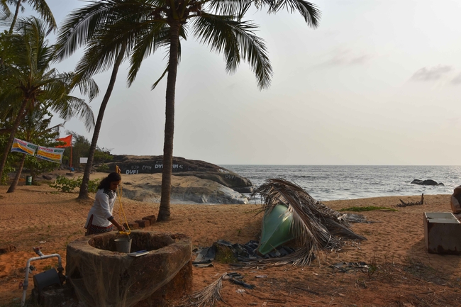 A sandy beach with tall palm trees beneath a hazy sky by the calm sea. In the foreground, a woman stands by a round stone fountain, lowering a metal bucket on a rope. Next to her lies an upturned green boat, partly covered with dry palm leaves. In the background, gentle waves break against dark rocks, and to the left, among the trees, several colourful signs and flags can be seen.