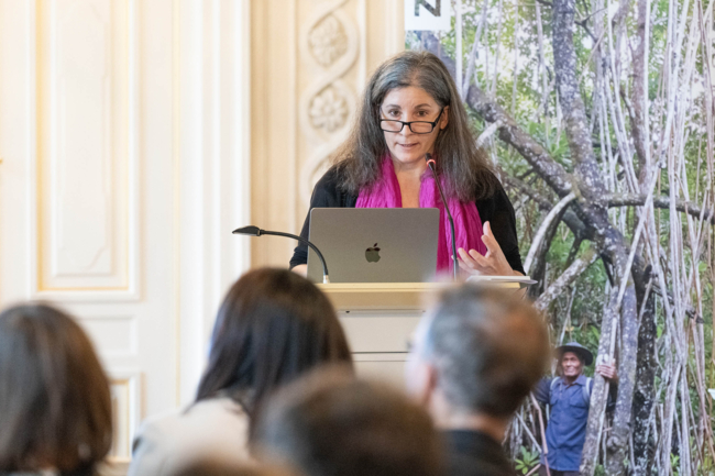 A woman with long dark hair stands at a lectern, in front of her is a laptop and behind her is a roll-up banner