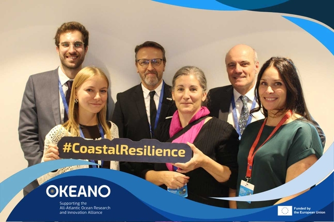 A group of six people, four men and two women, stand closely together and smile for a photo at an event. The woman in front holds a sign reading "#CoastalResilience" in bold yellow text.