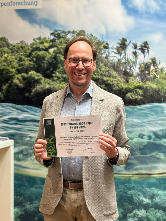 Ein Mann in einem hellen Anzug steht vor einer Wand mit einem Wellenfoto und hält ein Blatt mit der Aufschrift "Most Downloaded Paper in der Hand", er lächelt in die Kamera