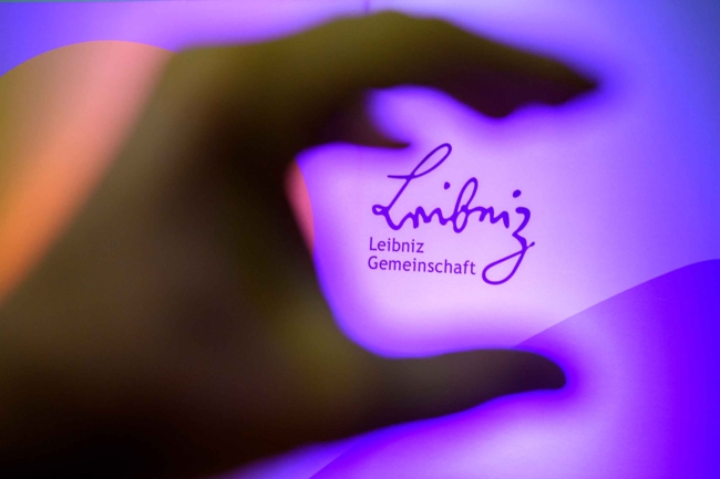 ChatGPT:  A close-up photograph shows a dark, out-of-focus hand in the foreground forming a loose "C" shape with the fingers and thumb. The background features a glowing purple and pink gradient with the "Leibniz Gemeinschaft" logo in elegant, cursive script.