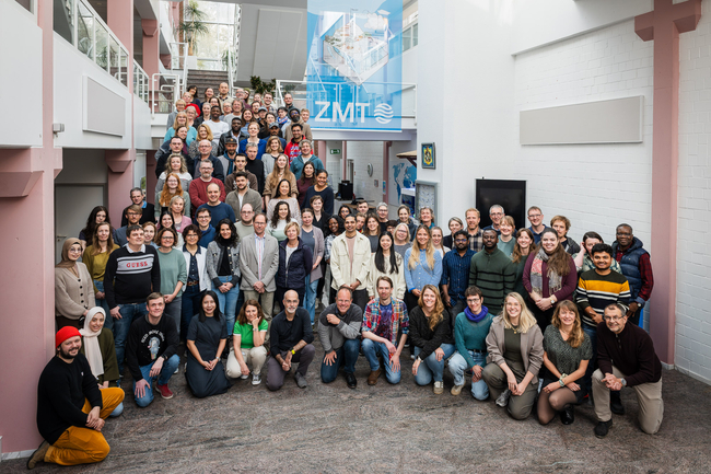 The photo shows a large group of people who have assembeld in a lightly lit foyer od a building. They are smiling for the camera.