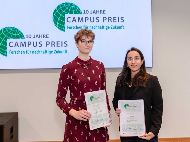 Zwei Frauen stehen nebeneinander und halten Urkunden vor einem großen Bildschirm mit dem Logo „10 Jahre Campus Preis – Forschen für nachhaltige Zukunft“. Die Person links trägt eine Brille und ein bordeauxrotes Kleid mit weißem Vogelmuster, die Person rechts eine Brille, ein schwarzes Sakko und ein schwarzes Oberteil. Beide blicken in die Kamera und lächeln leicht in einer Veranstaltungslocation.