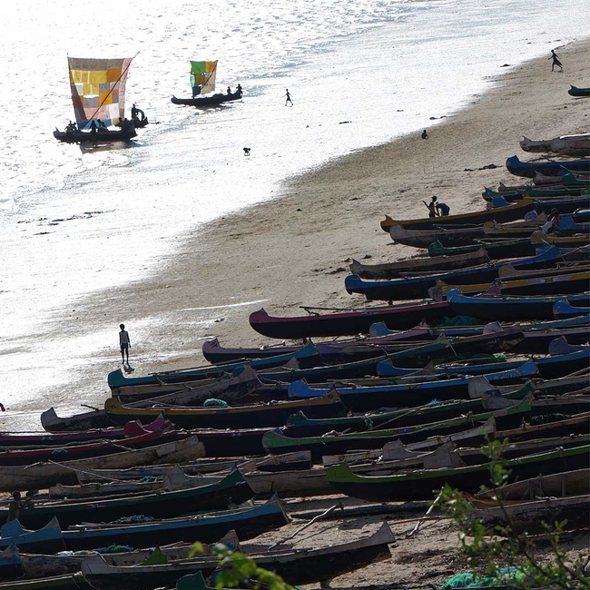Zahlreiche bunte Holzboote liegen am Sandstrand. Im flachen Wasser segeln kleine Boote mit farbigen Segeln, während einige Menschen am Ufer und im Wasser unterwegs sind. Das Sonnenlicht spiegelt sich auf der Meeresoberfläche.