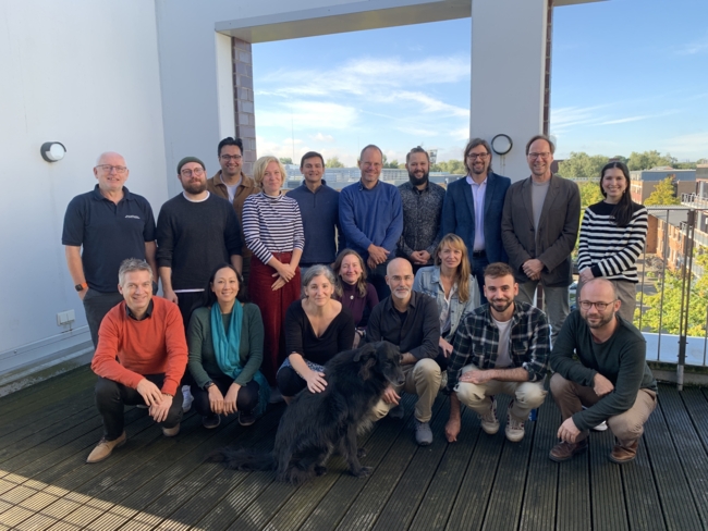 Group photo taken outdoors on a rooftop terrace. The image features 17 people, standing and sitting in two rows, smiling at the camera. In the front, there is a black dog sitting among the group. The background shows a clear blue sky and some distant buildings. The individuals appear to be participating in a workshop or meeting on a sunny day.